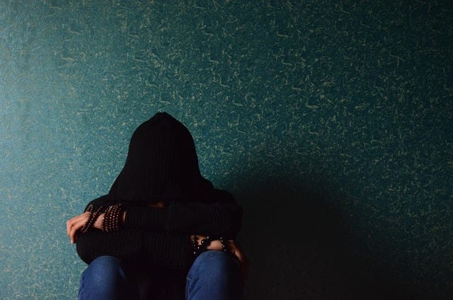 Sad woman sitting against a wall with head on her arms, face covered by a hoodie.