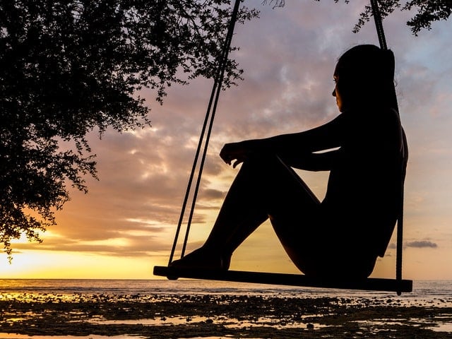Why does the dark night of the soul occur? Silhouetted image of young woman sitting on a swing looking out at a sunset.