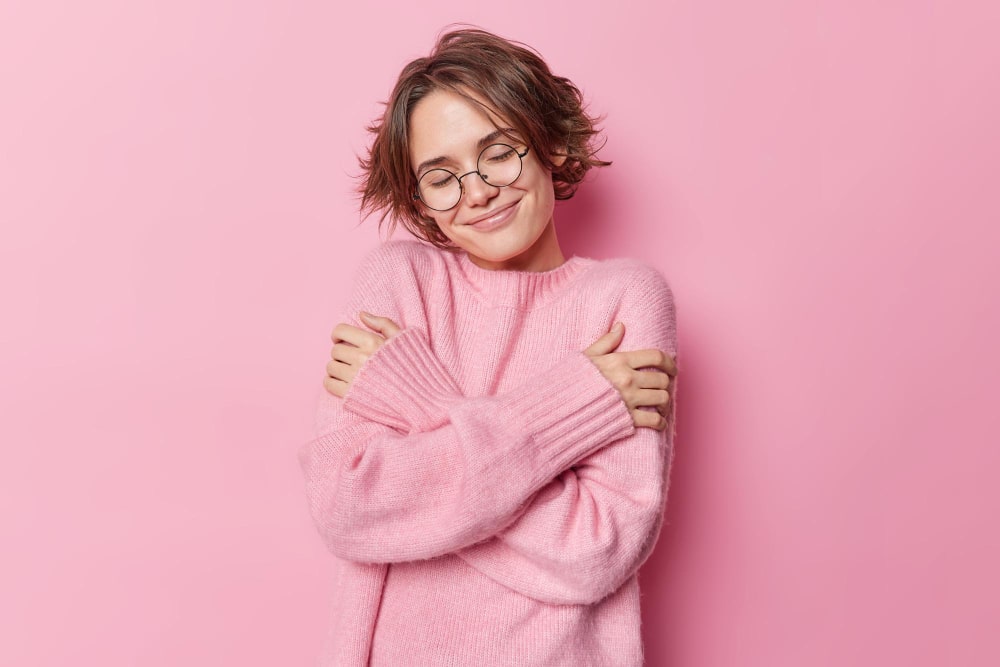 Radical self acceptance: woman in a pink jumper against a pink background hugging herself and smiling.